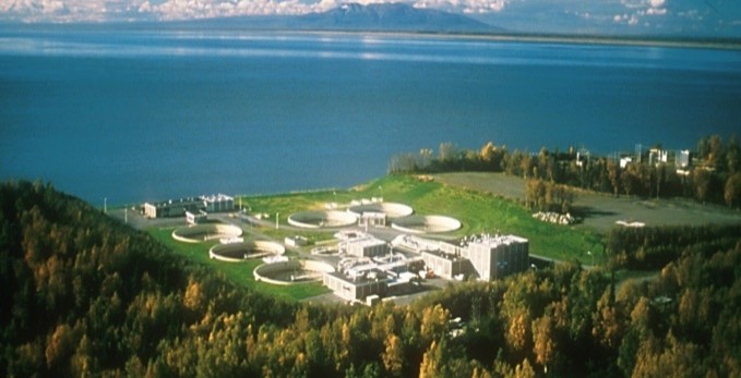Aerial view of the John M. Asplund Wastewater Treatment Facility, showing multiple circular treatment basins and operational buildings surrounded by forested land along a large body of water, with distant mountains and clouds on the horizon.
